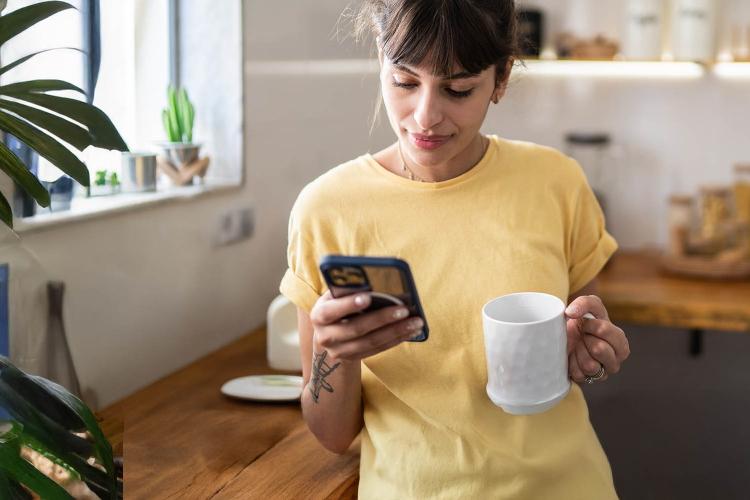 Frau mit Kaffeetasse in der Hand schaut in der Küche auf Smartphone
