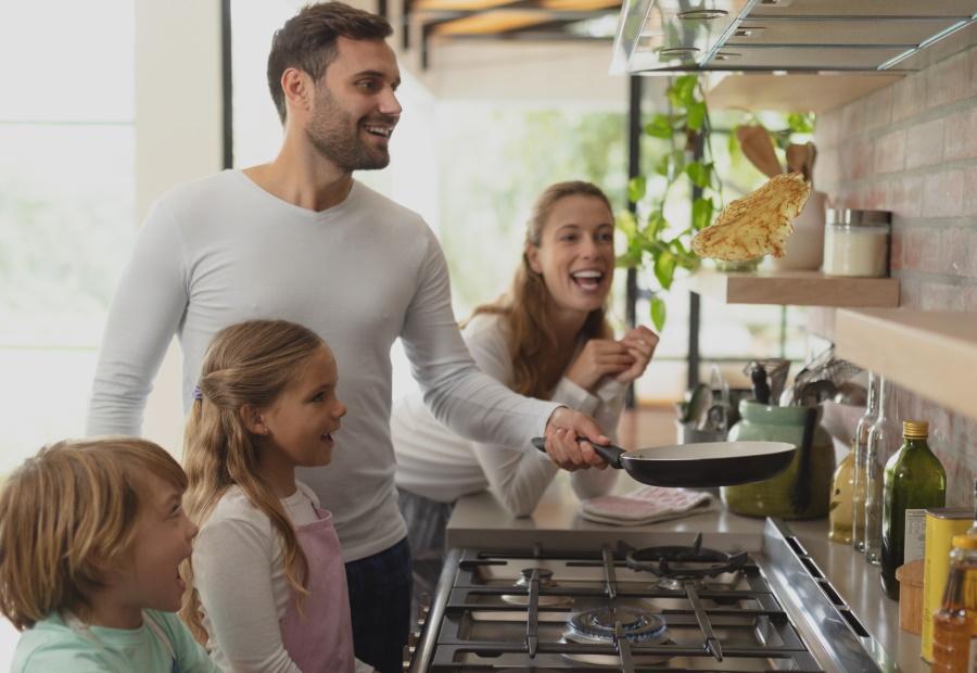 Familie steht am Gasherd, der Vater wirft einen Pfannkuchen aus der Pfanne durch die Luft