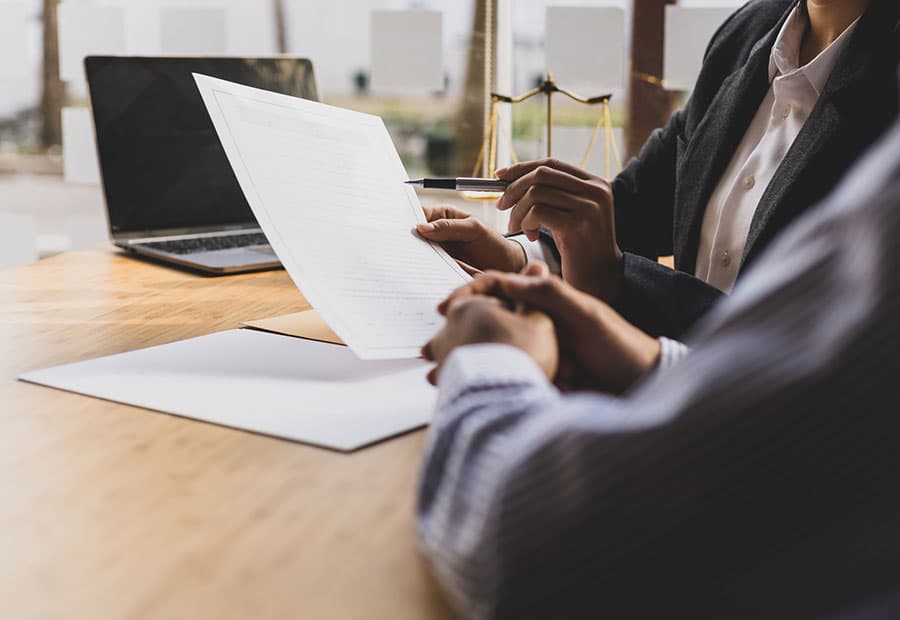 Zwei Personen sitzen an einem Tisch, eine hält ein Blatt Papier und zeigt mit einem Stift darauf, im Hintergrund ein Laptop und eine kleine Waage