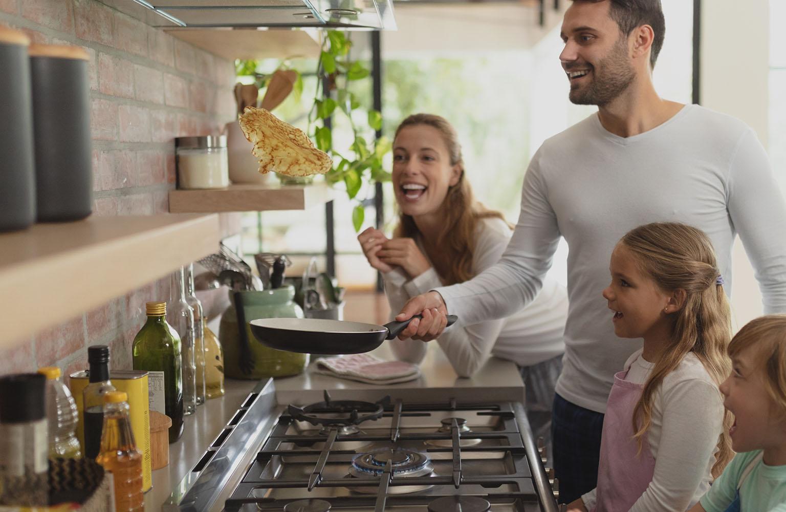 Familie steht am Gasherd, der Vater wirft einen Pfannkuchen aus der Pfanne durch die Luft