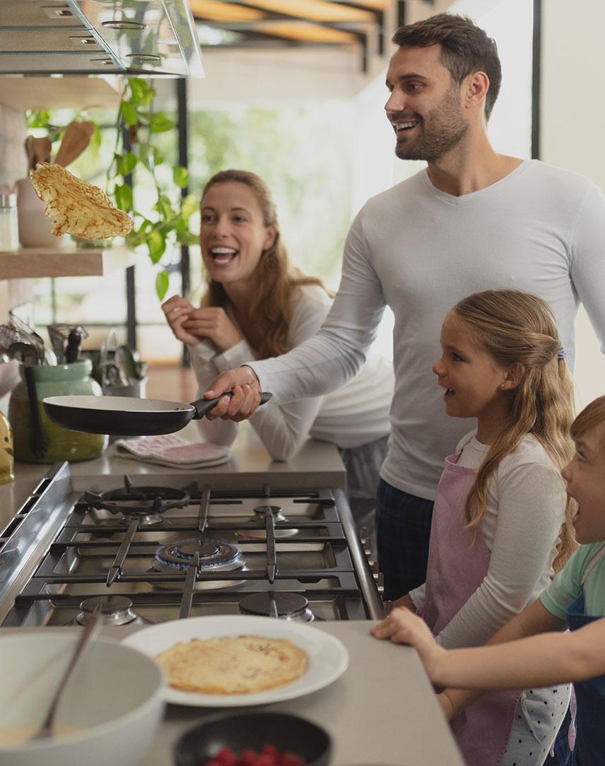 Familie steht am Gasherd, der Vater wirft einen Pfannkuchen aus der Pfanne durch die Luft