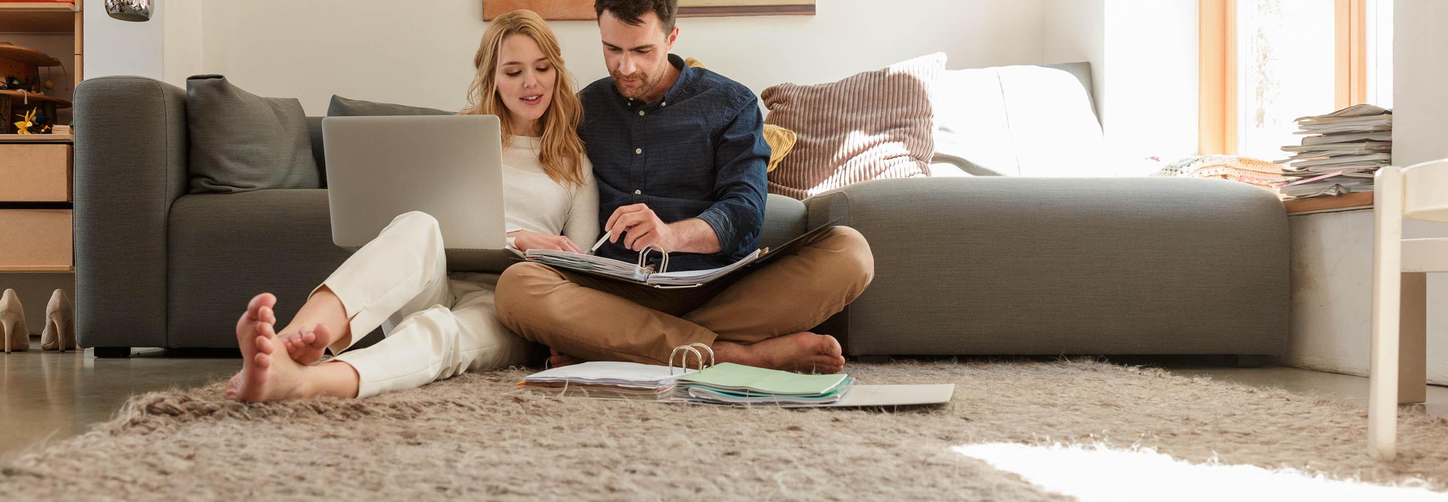 Ein junges Paar sitzt mit Aktenordner und Laptop auf dem Boden vor der Couch