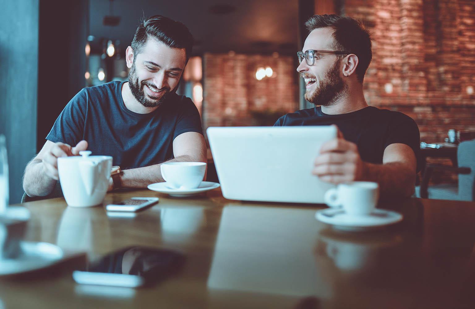 Zwei Männer im Café lachend am Laptop.