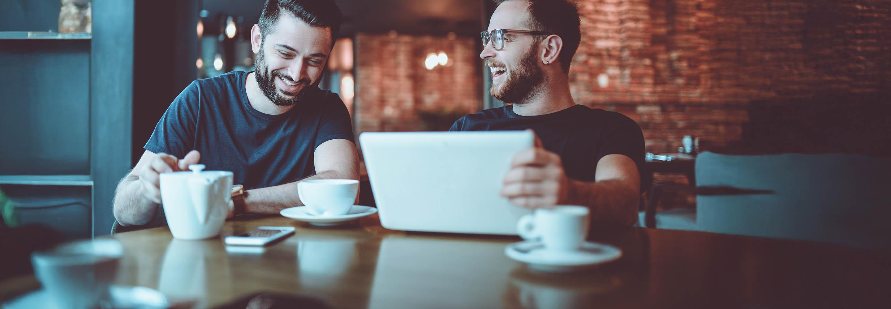 Zwei Männer im Café lachend am Laptop.