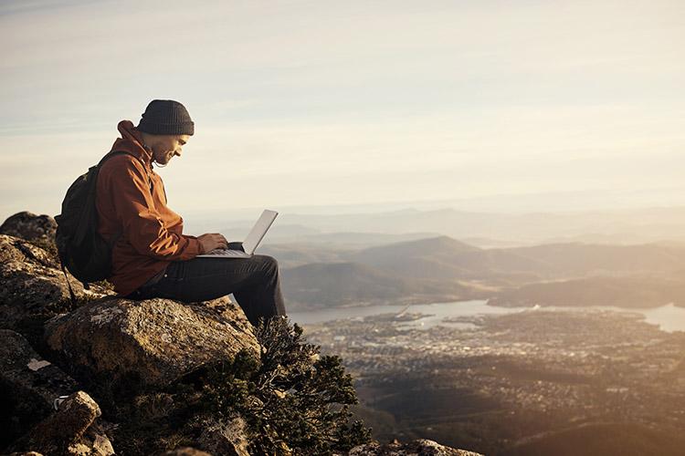 Mann sitzt in den Bergen mit Laptop auf dem Schoß und Smartphone in der Hand.