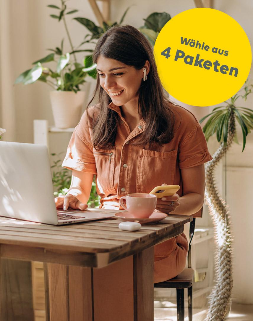 Junge Frau schaut lächelnd auf ihren Laptop bzw. ihr Handy, während sie mit Kopfhörern im Ohr telefoniert