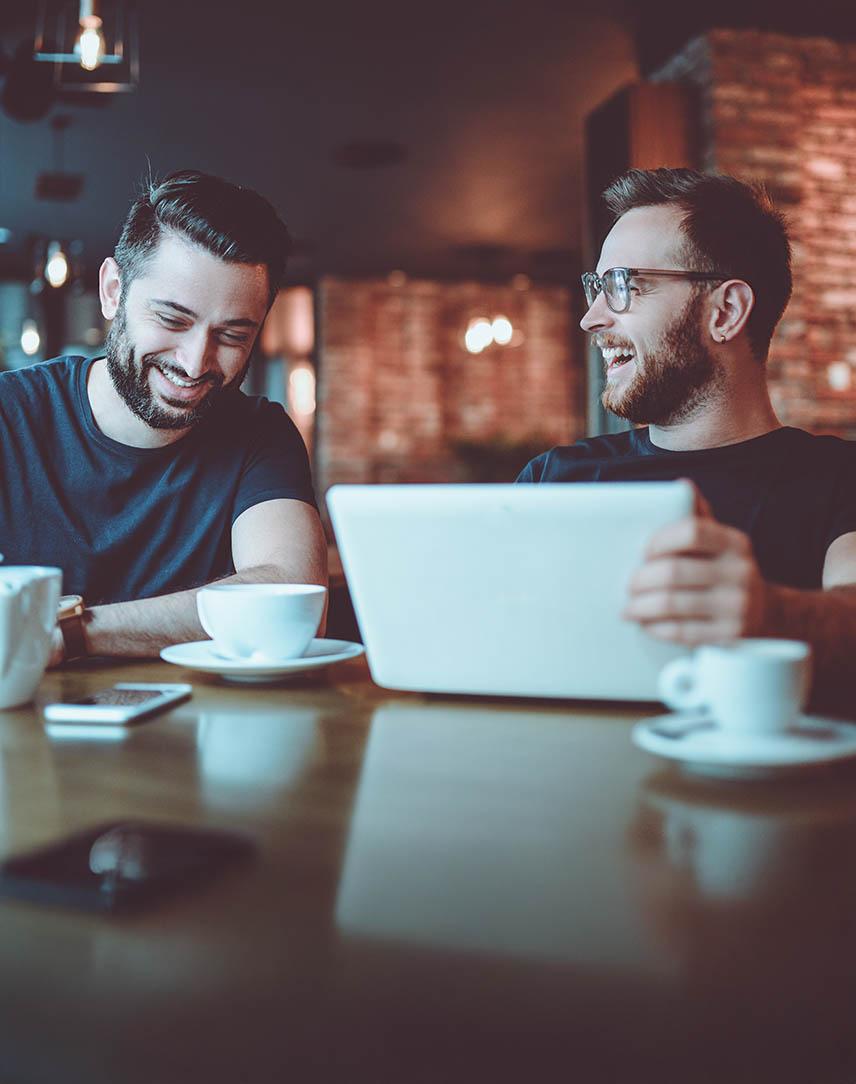Zwei Männer im Café lachend am Laptop.